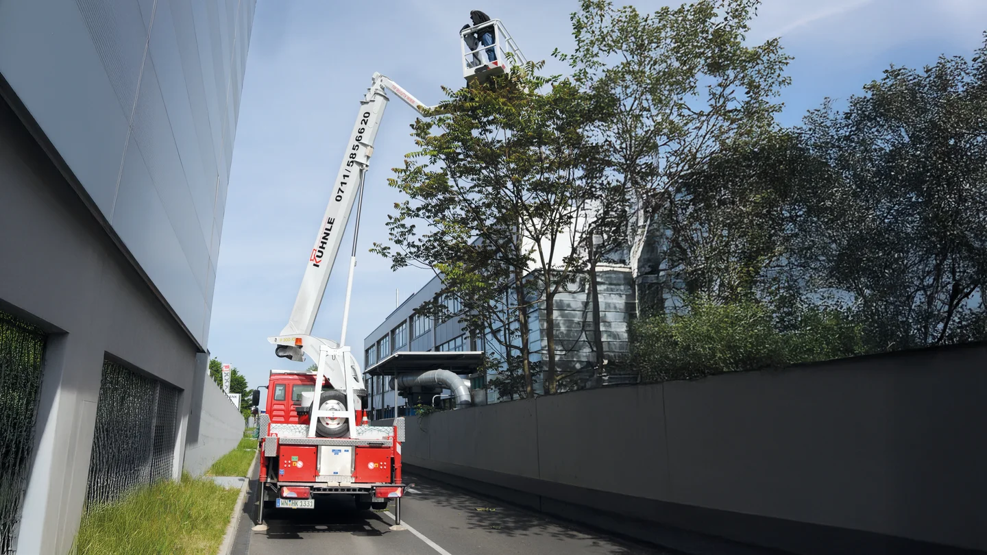 Rote ausgefahrene Arbeitsbühne zwischen zwei gläsernen Gebäuden, rechts ein Baum.