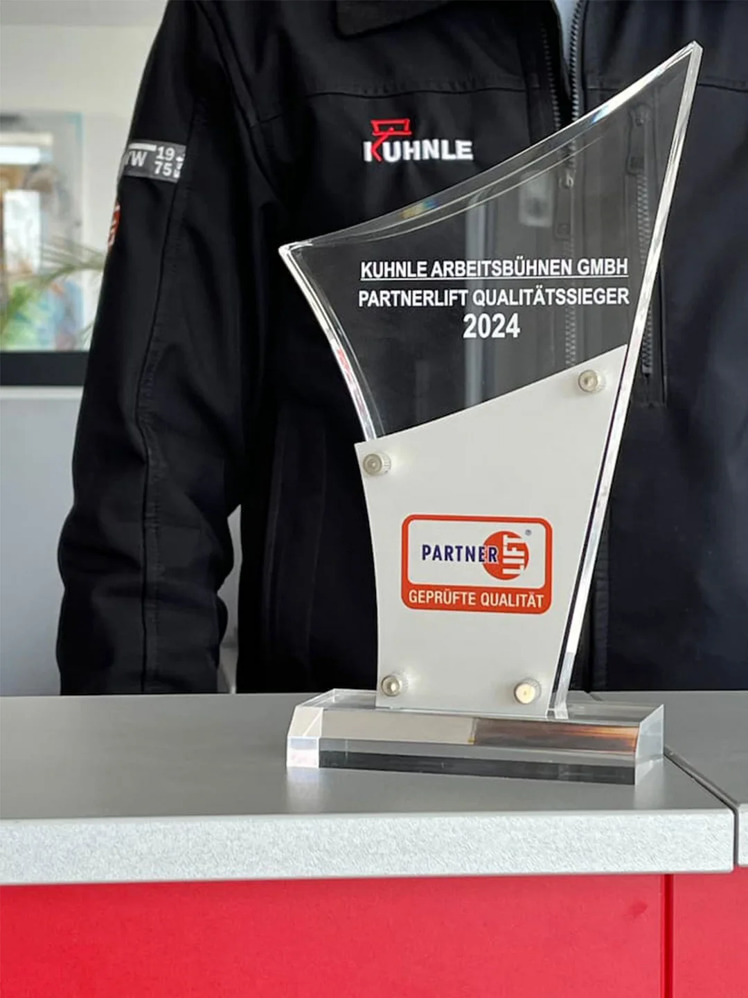 Award auf Tisch, dahinter Person in schwarzer Arbeitsjacke.