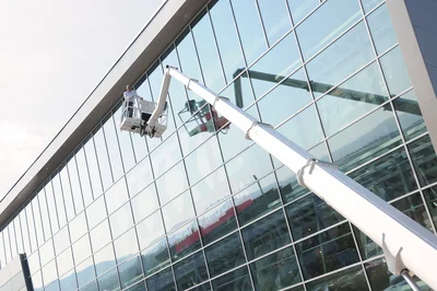 Ausgefahrene Arbeitsbühne vor hohem gläsernem Gebäude, darauf eine Person, links oben Himmel und Wolken.