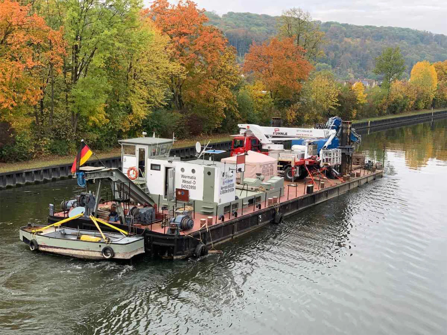 Boot auf Wasser, darauf Arbeitsbühnen; hinten Bäume.
