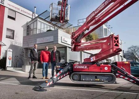 Gebäude, davor drei Personen, rechts rote Arbeitsbühne.
