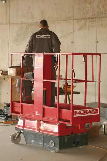 Person auf roter Arbeitsbühne auf dem Boden, in Arbeitsumgebung mit Schubkarre, hinten eine Steinwand.