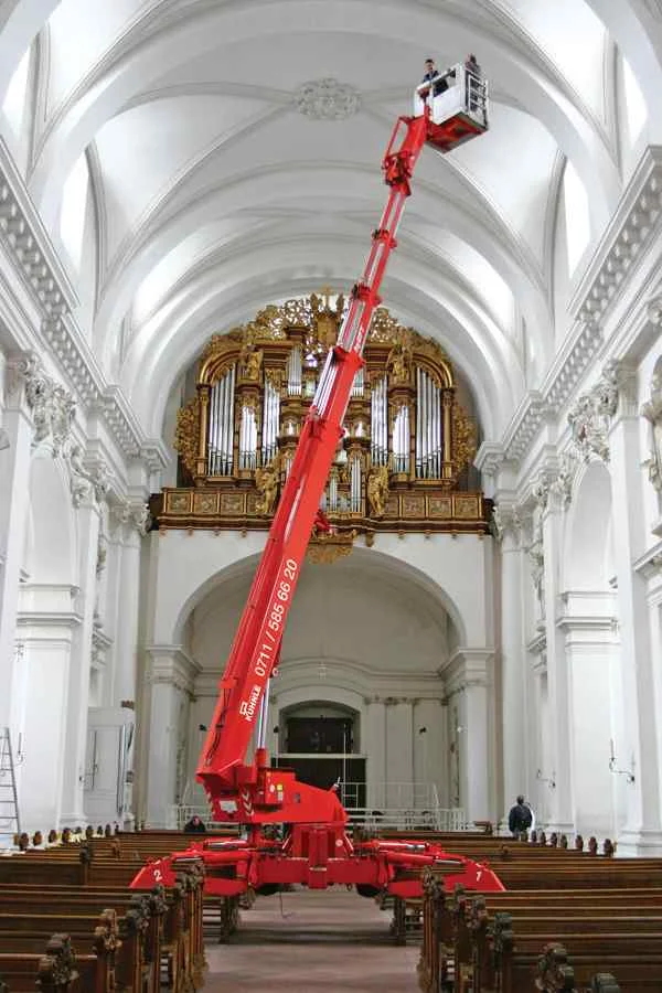 Ausgefahrene Arbeitsbühne mittig in einer Kirche, davor Sitzbänke.