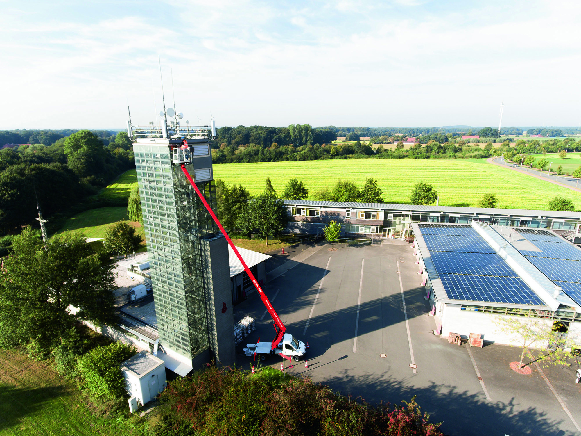 Vogelperspektive mit hohem Gebäude auf großem Hof und Arbeitsbühne links, niedrigem weißen Gebäude rechts sowie Feld und Wald im Hintergrund.